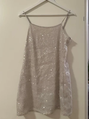  Paljettklänning - Säljer en glittrig champagnefärgad kortklänning med tunna axelband. Klänningen har ett enkelt, rakt snitt och är täckt av paljetter som ger ett snyggt skimmer. 