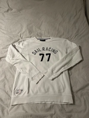 Vit Sail Racing sweatshirt med tryck - Vit långärmad sweatshirt från Sail Racing med svart tryck 'SAIL RACING 77' på bröstet och en patch med logga vid nederkanten. Rund hals och mjukt material, perfekt för chill dagar. Klassisk och sportig stil med normal passform. Finns fläckar se bild!