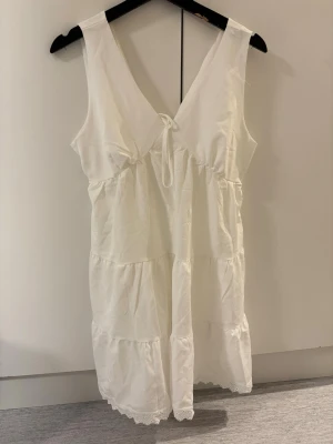 Vit ärmlös klänning med knyt - Säljer en vit, ärmlös klänning i storlek L med v-ringning och knyt framtill. Klänningen har volangdetaljer och spetskant längst ner.