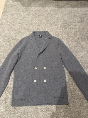 Italiensk grå stickad blazer - Snygg grå kavaj med dubbelknäppning och ljusa knappar från Andrea Fenzi. Jackan har två stora fickor framtill och klassisk krage. Materialet är 100% merinoull, känns mjuk och lent. Blazern är Made In Italy. 