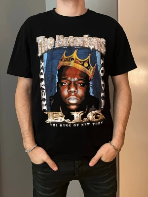 Svart Notorious B.I.G. t-shirt - Svart t-shirt med stor färgglad print framtill med The Notorious B.I.G.-motiv och texten 'Ready to Die' och 'The King of New York'. Klassisk passform med rund hals och korta ärmar. Perfekt för dig som gillar hiphop och streetwear.