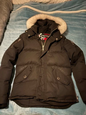 Svart Moose Knuckles dunjacka med päls - Säljer en svart dunjacka från Moose Knuckles med fluffig ljus päls på huvan. Jackan har långa ärmar, dragkedja, knappar och två fickor framtill. Märkets logga sitter på ärmen och insidan har ett broderat hockeymotiv. Perfekt för kalla dagar!