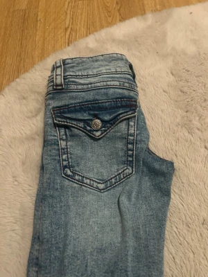Bootcut jeans från Gina Tricot, blå - Snygga ljusblå bootcut jeans från Gina Tricot i storlek 140. Jeansen har slitna detaljer på benen, klassiska fickor med knapp och röda sömmar som kontrast. Perfekt för dig som gillar en avslappnad och trendig look.