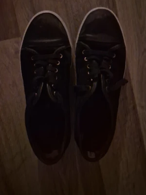 Svarta sneakers från Lanvin - Säljer ett par klassiska svarta sneakers från Lanvin med vita sulor och svarta skosnören. Skorna har rund tå, metallöljetter och en stilren design som passar till det mesta. Perfekta för dig som gillar en clean och tidlös look.