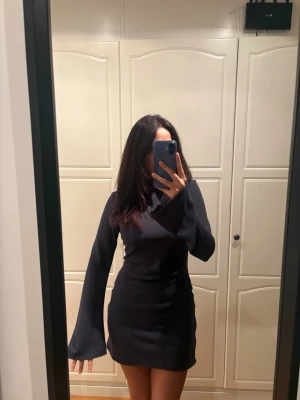 Mörkblå kort klänning från H&M - Säljer en mörkblå kort klänning från H&M med långa vida ärmar och öppen rygg med en söt rosettdetalj, i storlek XS. Den är i mycket bra skick då den bara använts en gång. 