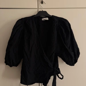 Svart omlottblus med puffärm - Säljer en svart omlottblus med puffiga trekvartsärmar och knytband i midjan. Blusen har en v-ringad design och är tillverkad i ett lätt, krinklat tyg som ger en cool struktur. Perfekt för dig som gillar stilrena och trendiga plagg.