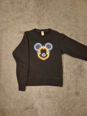 Svart Disneytröja Uniqlo  - Cool svart sweatshirt från Uniqlo x Disney i storlek XS. Framsidan har ett neoninspirerat tryck av Musse Piggs ansikte i blått, vitt och gult. Tröjan har rund halsringning och långa ärmar. Perfekt för dig som gillar streetstyle och Disney.