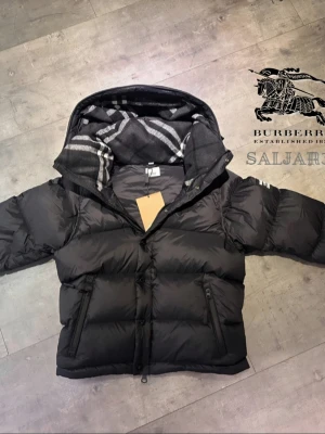 Burberry Pufferjacka  - Svart dunjacka från Burberry med klassisk Säljer en elegant och mångsidig Burberry-jacka i svart, i nyskick. Dunfodrad för värme och komfort med det klassiska rutmönstret i huvan som ger en exklusiv touch. Praktiska dragkedjefickor, avtagbar huva och diskret logopatch på ärmen. Jackan är i princip som ny och utan defekter. En lyxig kombination av stil och funktion – perfekt för både vardag och tillfällen då du vill klä upp dig.  📦Snabb leverans - skickas samma dag (om ej hindr)📌..