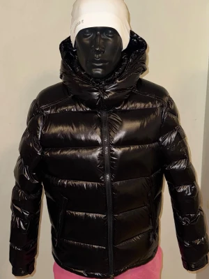 Svart pufferjacka från Moncler - Säljer en svart, glansig pufferjacka från Moncler med huva och dragkedja. Jackan har en ficka med Moncler-logga på ärmen och quiltad design. Perfekt för kalla dagar och riktigt snygg streetstil.