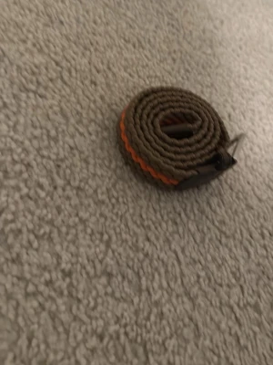 Brun och orange flätad bälte - Säljer ett snyggt flätat bälte i brunt med en orange detalj längs hela bandet. Bältet har ett metallspänne och är gjort i ett slitstarkt syntetmaterial. Perfekt för att ge outfiten lite extra färg och stil.