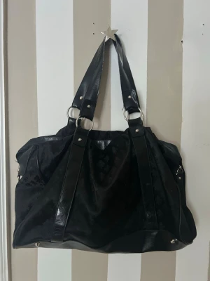 Stor svart axelväska - Säljer en stor svart axelväska med snyggt mönster och breda axelremmar i skinnimitation. Väskan har silverfärgade ringdetaljer och dragkedja upptill. Perfekt för dig som vill ha plats för allt.