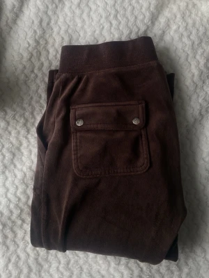 Bruna mjukisbyxor Juicy Couture XS - Säljer ett par bruna mjukisbyxor från Juicy Couture i storlek XS. Byxorna har bred resår i midjan med snörning, stora fickor fram och bak med metallknappar och ett litet broderat JC-logo på ena benet. Mjukt material som är skönt att ha på sig.