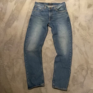 Blå jeans från Nudie Jeans Co W30 L32 - Säljer ett par klassiska blå jeans från Nudie Jeans Co i storlek W30 L32. Jeansen har fem fickor, orangea sömmar och Nudie-logga på bakfickan. Modellen har rak passform och är tillverkad i slitstarkt bomullsmaterial. Perfekt för dig som gillar tidlös denimstil.