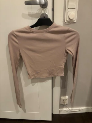 Beige croppad långärmad topp - Säljer en beige croppad topp med lång ärm och rund hals. Toppen är enkel och stilren, tillverkad i mjukt bomullsmaterial med en lätt ribbad struktur. Perfekt att matcha med jeans eller kjol för en chill look.