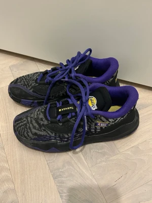 Svarta och lila Tarmak Lakers sneakers 36 - Säljer ett par Tarmak Lakers sneakers med svart och grått mönster, lila detaljer och snörning. Lakers-logga på plösen och NBA-märke på sidan. Insidan är lila på kanten och gul innersula, och sulan under har ett unikt mönster i svart och lila. Texten 'PURPLE' och 'GOLD' bak på hälen. Storlek 36 och endast använda fåtal gånger.