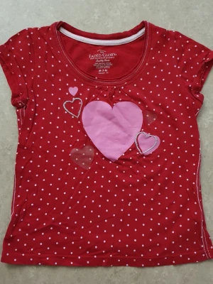 T-shirt - Säljer en röd t-shirt från Faded Glory med vita prickar och flera hjärtan i rosa och glitter på framsidan. T-shirten har rund hals och korta ärmar, perfekt för dig som gillar färgglada och söta prints. Materialet är mjuk bomull.