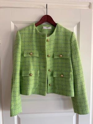 Grön tweed kavaj från Mango L oanvänd  - Snygg grön kavaj från Mango i tweed med gul och lila inslag. Modellen är croppad med fyra fickor framtill och guldfärgade knappar. Jackan har rund hals och är helfodrad med ljusgrönt tyg. Perfekt för dig som vill sticka ut med färg.