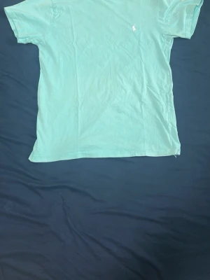 Turkos t-shirt från Polo Ralph Lauren - Säljer en fräsch turkos t-shirt från Polo Ralph Lauren med klassisk vit logga broderad på bröstet. T-shirten har korta ärmar och är gjord i mjuk bomull som känns skön mot huden. Perfekt för dig som gillar stilrena och enkla plagg.