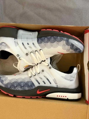 Nike Presto sneakers med mönster - Nike Presto sneakers i vitt med svart och rött. Skorna har ett unikt cirkelmönster i blått på ovandelen, röd swoosh och svart sula med röda detaljer. Ovandelen är i syntet och mesh, med snörning och rund tå. Perfekt för dig som gillar coola och bekväma sneakers.