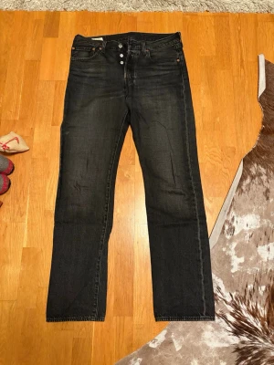 Levi's 501 mörkgrå jeans W32 L34 - Mörkgrå Levi's 501 jeans med klassisk rak passform och knappgylf. Jeansen har fem fickor och är tillverkade i slitstarkt denim. Perfekt för dig som gillar tidlös stil och vill ha ett par jeans som funkar till allt. Märke på bakfickan och läderpatch i midjan.