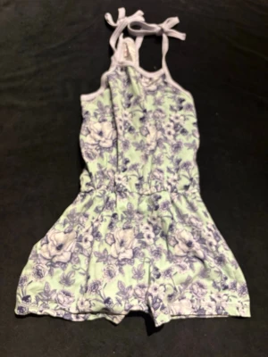 Blommig byxdress med knytband - Säljer en söt byxdress med ljuslila och vit blommönster på ljusgrön botten. Byxdressen har knytband vid axlarna och mjuka kanter. Perfekt för dig som gillar unika plagg med blommig vibe och bekväm passform.