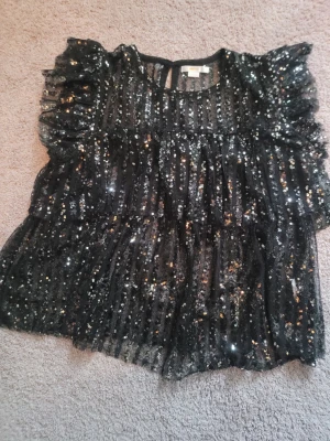 Svart paljettblus från Gina Tricot - Säljer en svart blus från Gina Tricot med glittriga paljetter över hela toppen. Blusen har voluminösa korta ärmar och är tillverkad i ett tunt, transparent syntetmaterial. Perfekt för att sticka ut med extra glitter och volym.