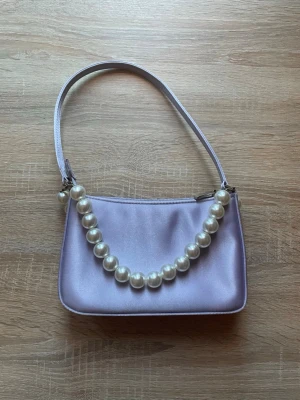 Ljuslila axelväska från Barratts - Snygg ljuslila axelväska från Barratts i satinliknande material. Väskan har en enkel, stilren design med dragkedja upptill och ett innerfack med dragkedja. Perfekt storlek för det viktigaste och en riktigt trendig accessoar. Två avtagningsbara pärlband ingår.