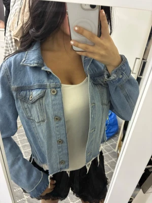 Ljusblå croppad jeansjacka - Säljer en ljusblå croppad jeansjacka med rå kant nertill och klassiska bröstfickor med knappar. Jackan har långa ärmar och en avslappnad passform. Perfekt att slänga över en topp för en chill look.