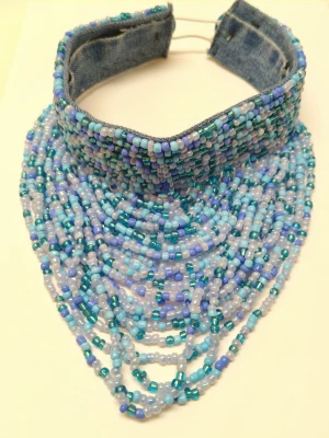 Pärlat halsband i blå toner - Unikt halsband med denimkrage täckt av små pärlor i olika blå och turkosa nyanser. Halsbandet har ett halsnära snitt och flera lager av pärlor som hänger ner över bröstet. Perfekt för dig som vill sticka ut med ett statement smycke.
