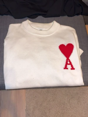 Vit stickad tröja från Ami - Säljer en vit stickad tröja från Ami Alexandre Mattiussi med röd hjärtlogga och bokstaven A på bröstet. Tröjan har rund halsringning, ribbade muddar och lång ärm. Perfekt för dig som gillar stilrena plagg med en twist.