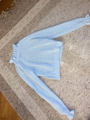 Ljusblå stickad tröja med volang - Supersöt ljusblå stickad tröja från H&M Divided i storlek S. Tröjan har långa ärmar och volangdetaljer vid halsringningen och ärmsluten som ger en extra gullig touch. Perfekt att styla med jeans eller kjol för en chill look.