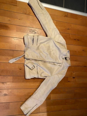 Beige mocka bikerjacka från Zara - Säljer en beige bikerjacka i mocka från Zara. Jackan har flera silvriga dragkedjor, bälte i midjan och en cool asymmetrisk dragkedja framtill. Ståkrage med knapp och långa ärmar med dragkedjor vid ärmsluten. Perfekt för dig som vill ha en edgy look.