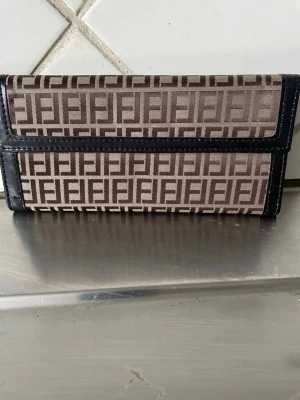 Fendi plånbok med FF-mönster - Säljer en snygg plånbok från Fendi med det klassiska FF-mönstret i brunt och beige. Plånboken har svarta skinnkanter och flera fack för kort och ID. Perfekt för dig som gillar designer och vill ha något stilrent och praktiskt.