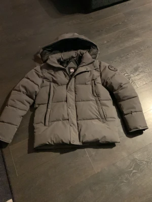 Grå pufferjacka från Canada Goose - Säljer en grå pufferjacka från Canada Goose med huva och flera fickor framtill. Jackan har en tydlig logotyp på ärmen och är fodrad för extra värme. Perfekt för kalla dagar och har en stilren look som funkar till det mesta.