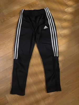 Svarta Adidas träningsbyxor med dragkedja - Svarta träningsbyxor från Adidas med klassiska vita ränder längs sidorna och logga på låret. Byxorna har resår i midjan och dragkedjor vid bensluten för extra smidighet. Perfekta för sport eller chill. Tillverkade i polyester.