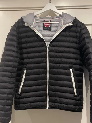 Svart pufferjacka från Colmar - Snygg svart pufferjacka från Colmar med vit dragkedja och huva. Jackan har quiltad design och två sidofickor med dragkedja. Insidan är ljusgrå och märket syns tydligt både på ärmen och insidan. Perfekt för kyliga dagar och riktigt stilren. Jackan har tvyärr två defekter som har lagats som man inte riktigt märker till 