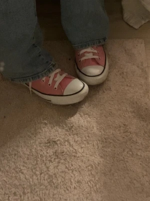 Rosa Converse Chuck Taylor All Star - Säljer ett par klassiska Converse Chuck Taylor All Star high tops i rosa med vita snören och tåhätta. Skorna har den ikoniska loggan på sidan, svart rand runt sulan och är tillverkade i slitstark bomullscanvas. Perfekta för dig som vill sticka ut lite extra!