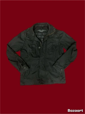 Svart fieldjacket från Hampton Republic - Svart fieldjacket från Hampton Republic med coola detaljer som bröstfickor med lock, knappstängning och dragkedja. Jackan har en klassisk krage och är tillverkad i bomullsmaterial. Perfekt för dig som gillar en stilren och avslappnad look.