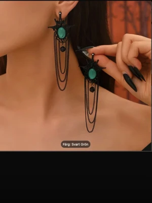 Svartgrön väst med kedjedetaljer - Cool väst i svart med gröna inslag, designad med flera tunna kedjor som hänger ner och en dekorativ grön oval detalj framtill. Västen har en unik form med spetsiga kanter och ger en edgy vibe. Perfekt för dig som vill sticka ut med din stil.