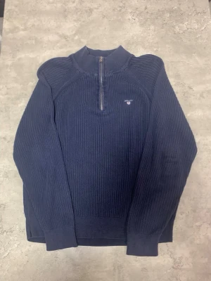 Marinblå stickad half zip från GANT - Säljer en marinblå stickad tröja från GANT med half zip och hög krage. Tröjan har ribbad struktur, lång ärm och GANT-logga broderad på bröstet. Perfekt för dig som gillar klassisk stil med en modern touch. 
