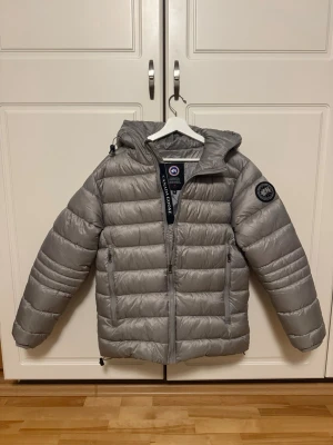 Ljusgrå dunjacka från Canada Goose - Hjälper min vän som säljer sin ljusgrå dunjacka från Canada Goose med huva och dragkedja framtill. Jackan har två sidofickor med dragkedja och en snygg patch på ärmen. Perfekt för kalla dagar och riktigt skön att bära. Jackan är i väldigt bra skick (intill nyskick) och är sparsamt använd. Om du har någon fråga eller vill se fler bilder är det bara att skriva!