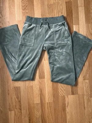 Juicy Couture mjukisbyxor XS olivgröna - Snygga olivgröna mjukisbyxor från Juicy Couture i storlek XS. Byxorna har bred resår i midjan med snörning, två stora fickor fram och två bak med knappar. Mjukt och glansigt material som känns lyxigt och chill. Perfekta för en avslappnad stil.