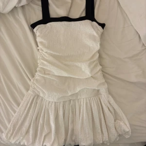 Vit kortklänning med svarta band - Supersöt vit kortklänning med svarta axelband och kant upptill. Klänningen har en rynkad, figurnära överdel och en volangig, puffig kjol. Materialet känns lätt och luftigt, perfekt för varma dagar. Stilren och trendig look som sticker ut.