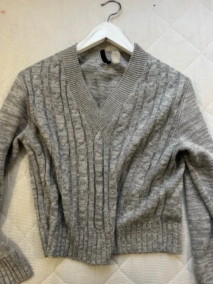 Grå kabelstickad tröja H&M - Säljer en grå kabelstickad tröja från H&M med v-ringad hals och ribbade muddar. Tröjan har lång ärm och klassisk kabelstickad struktur som ger en mysig och stilren look. Perfekt till höst och vinter för en chill vibe.