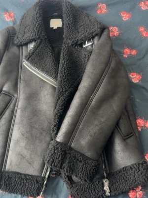 Svart teddyjacka från H&M XS - Säljer en svart teddyjacka från H&M i storlek XS. Jackan har grov teddyfoder, bred krage, detaljer med dragkedjor och spänne vid halsen. Utsidan är i syntetmaterial med matt finish och insidan är fluffig och mjuk. Perfekt för kalla dagar.