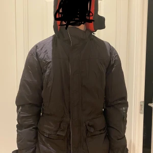 Svart vinterjacka från Jackgrossisten L - Säljer en svart vinterjacka från Jackgrossisten i storlek L. Jackan har röd och grå insida, hög krage och flera fickor. Perfekt för kalla dagar med sin vadderade och puffer-liknande design. Stängs med dragkedja och har justerbar midja.
