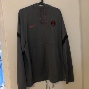 Grå PSG hoodie från Nike - Säljer en grå hoodie från Nike med Paris Saint-Germain-logga i svart och rött på bröstet. Tröjan har huva med snörning, lång ärm och en stor magficka. Snygga svarta detaljer på ärmarna och röd Nike-logga framtill. Perfekt för dig som gillar fotboll och streetwear.