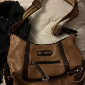 Betty Boop axelväska i brunt skinn -  axelväska från Betty Boop i brunt och mörkbrunt skinn med flera dragkedjefack och bred, randig axelrem. Insidan är fodrad med mönster av Betty Boop och hjärtan. Perfekt för dig som vill ha en unik och praktisk väska med snygga detaljer.