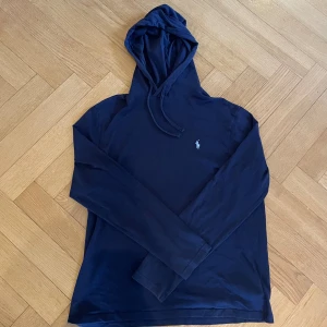Marinblå hoodie från Polo Ralph Lauren - Snygg marinblå hoodie från Polo Ralph Lauren med klassisk logga broderad på bröstet. Tröjan har huva med snörning och långa ärmar.