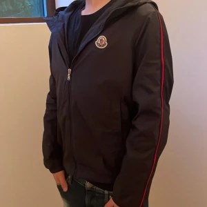 Svart vindjacka från Moncler med röd detalj - Svart vindjacka från Moncler med huva och dragkedja framtill. Snygg röd rand längs ärmarna och stort Moncler-tryck på ryggen. Jackan har en clean look och är perfekt för dig som gillar streetstyle. Materialet är lätt och känns sportigt. Köpte ny från nk 